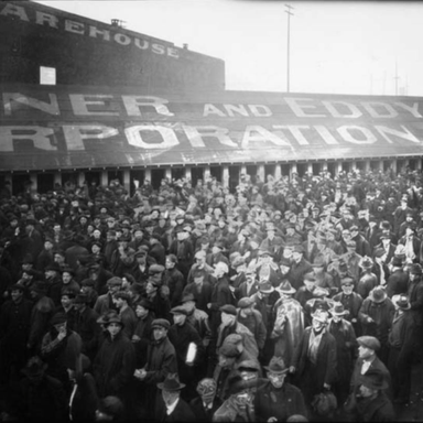 A black and white photo of a lot of men standing in line next to a warehouse